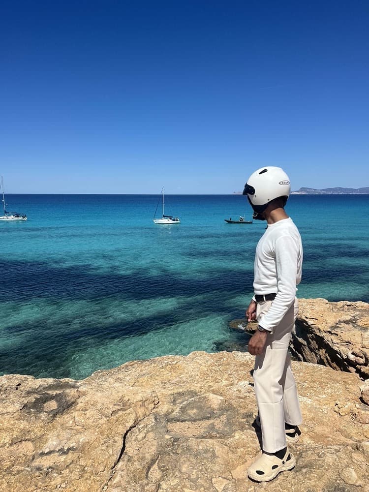 Jordan Gilbert in a white helmet on a rock ledge above clear turquoise water, sailing boats at anchor offshore.