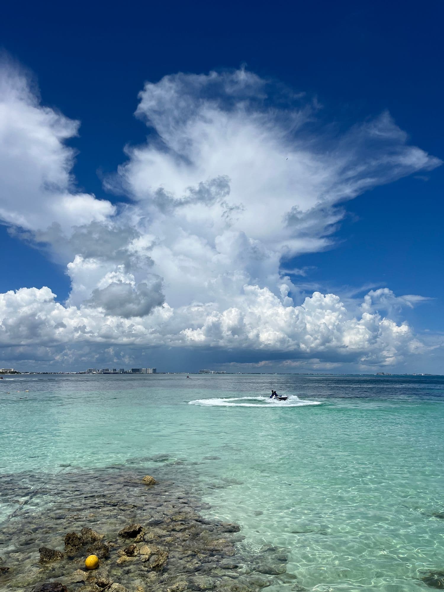 Turquoise shallows over rock below towering white clouds, a jet ski out on the water.
