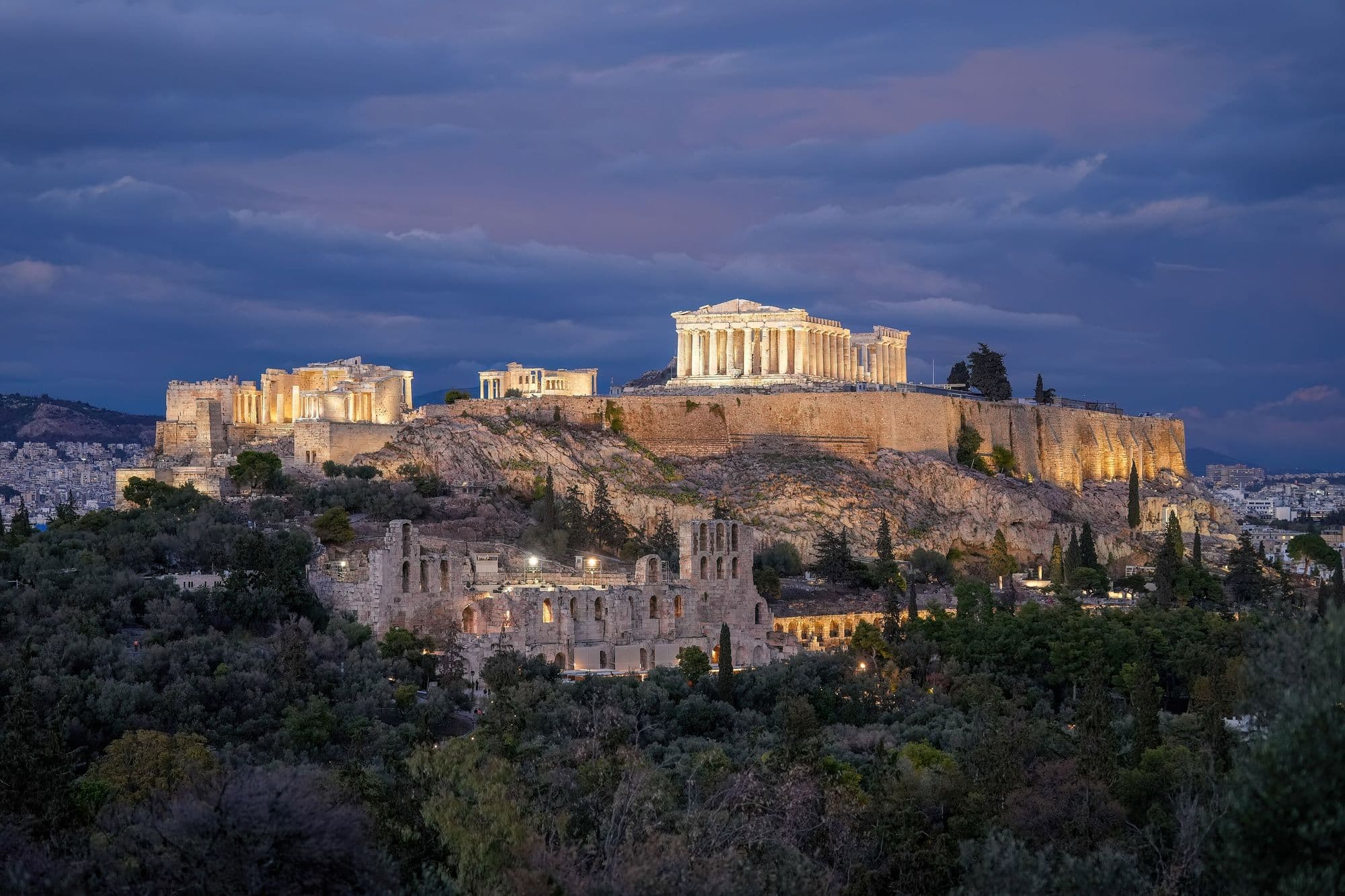 The Acropolis lit up at dusk on its hill above the trees, the Parthenon on top.