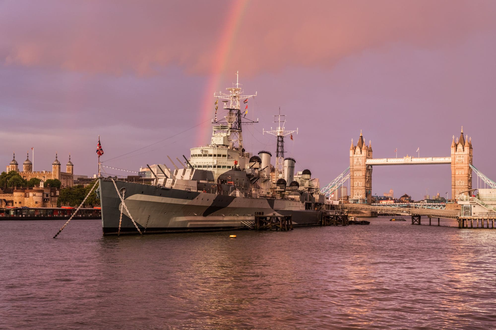 HMS Belfast moored on the Thames under a rainbow, with Tower Bridge and the Tower of London behind.