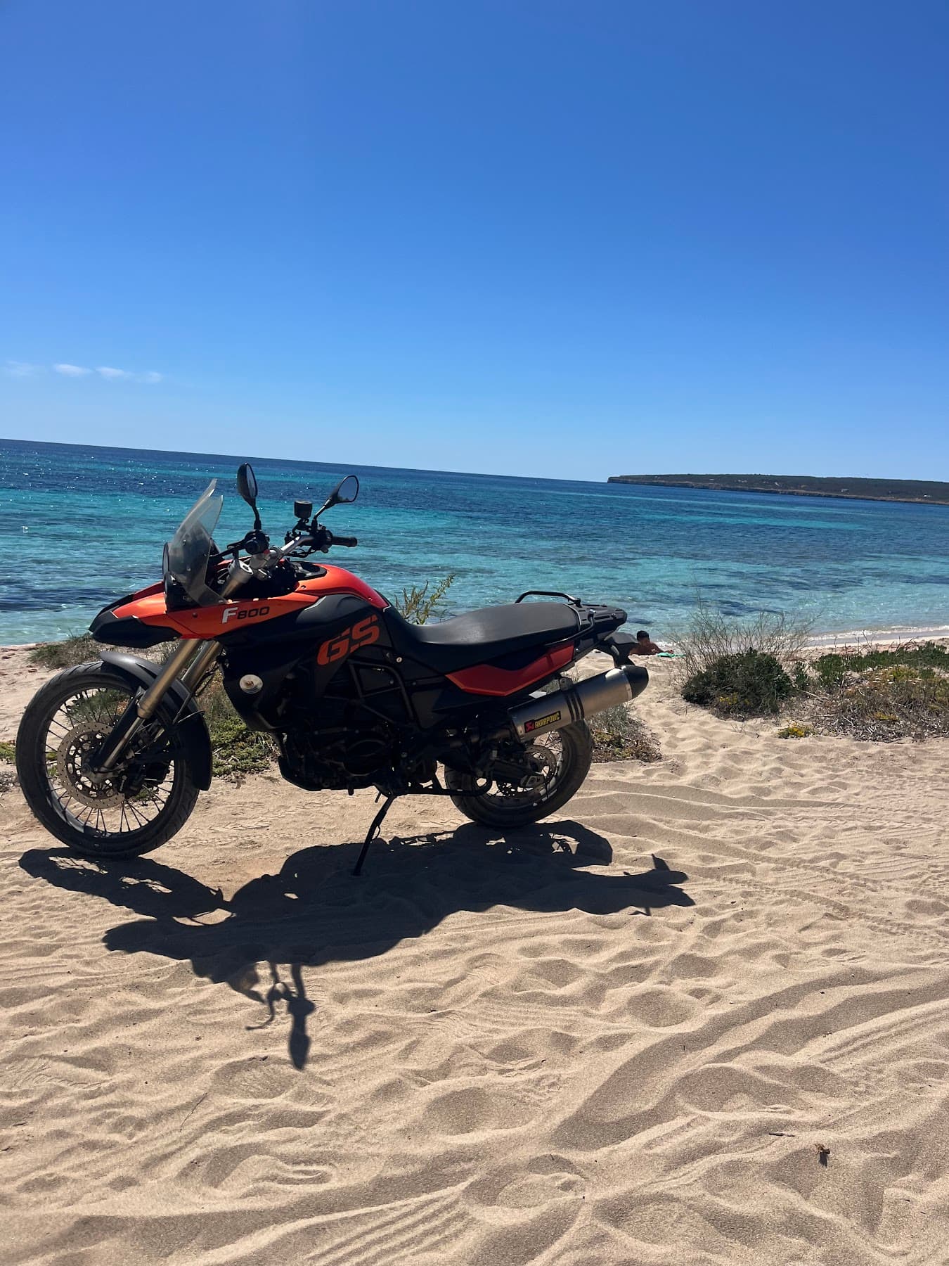 An orange adventure motorbike parked on a sandy track, the sea behind it.