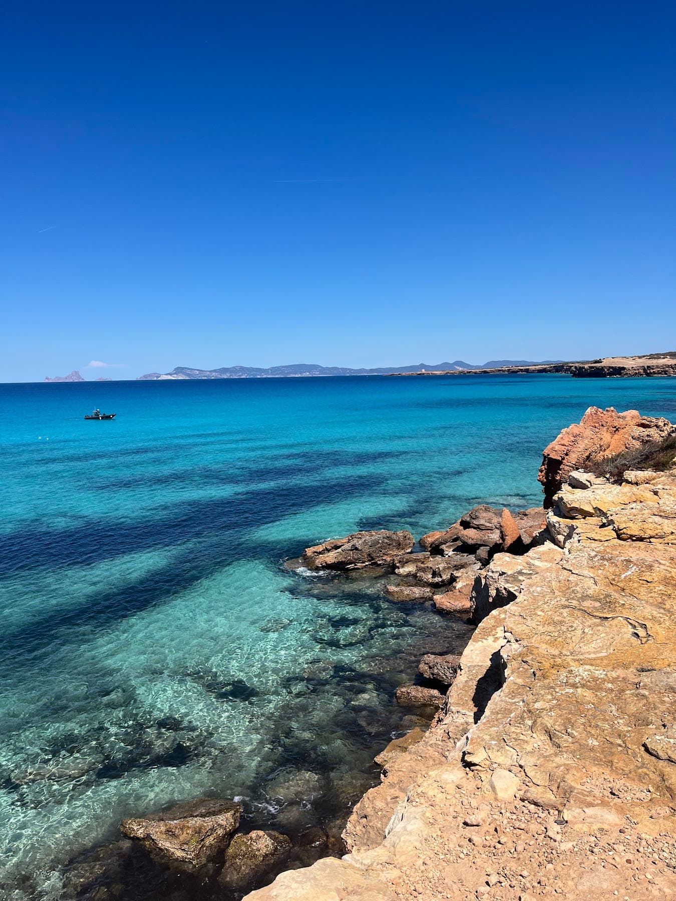 Clear turquoise water over rock and sand below a low cliff, hills on the horizon.