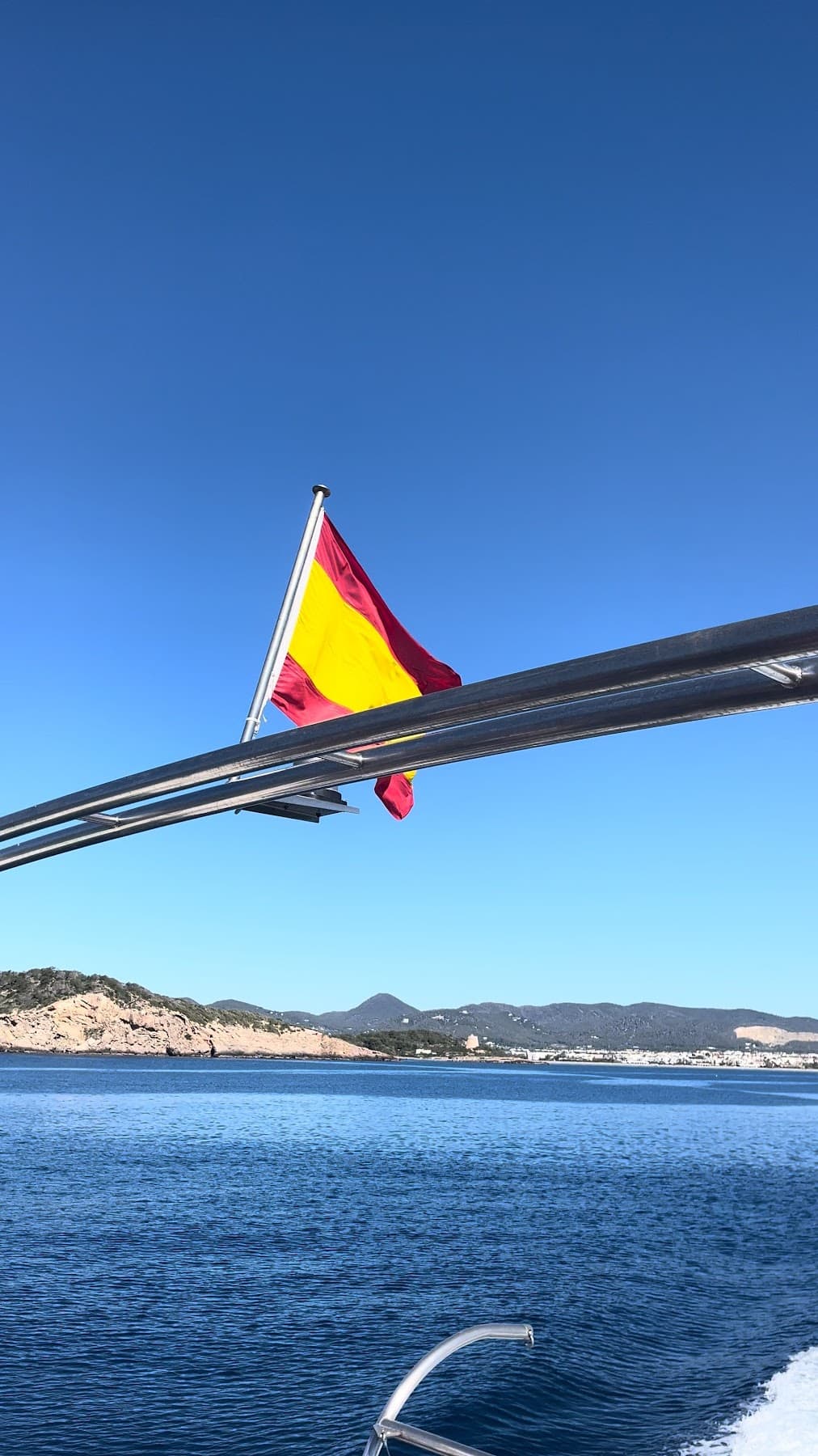 The Spanish flag on the stern rail of a boat at sea, with a coastline behind.